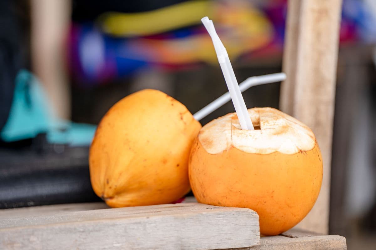 A glass of cool fresh coconut water with a straw next to a coconut