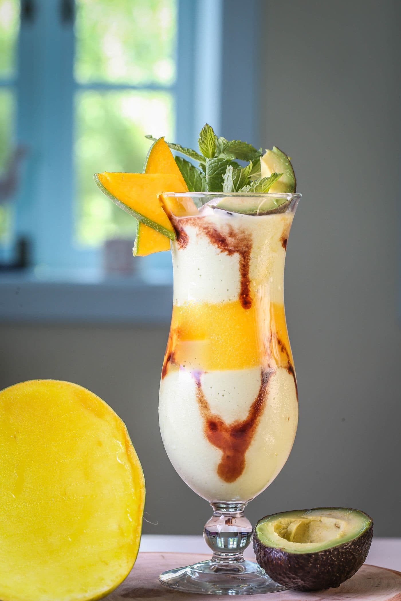 Ly cao sinh tố bơ xoài nhiều lớp trang trí lá bạc hà, miếng xoài và nửa quả bơ đặt cạnh trên bàn gỗ