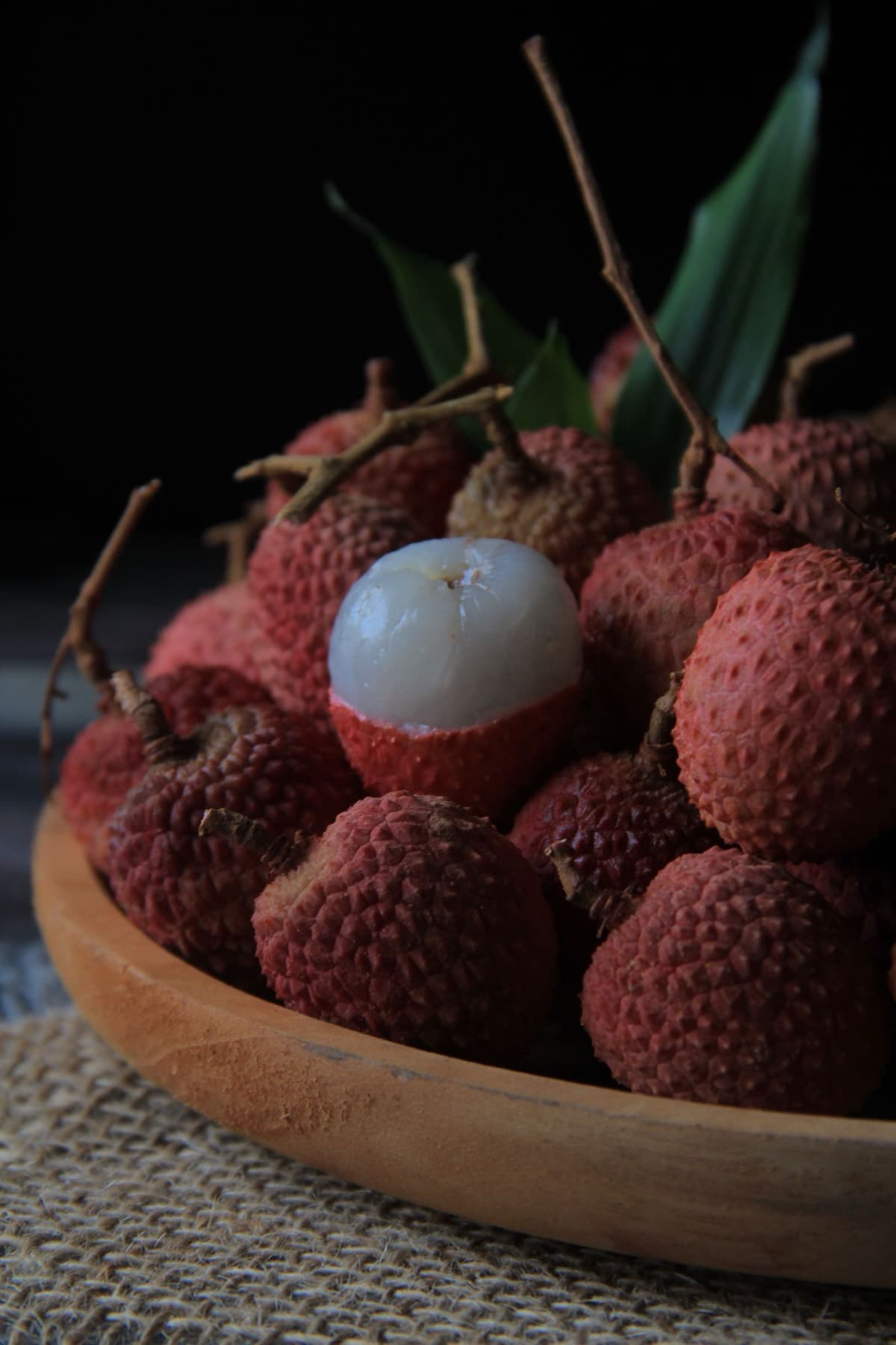 Đĩa gỗ đựng vải thiều tươi vỏ đỏ sần, một quả đã bóc vỏ lộ cùi vải trắng trong mọng nước, trên nền tối