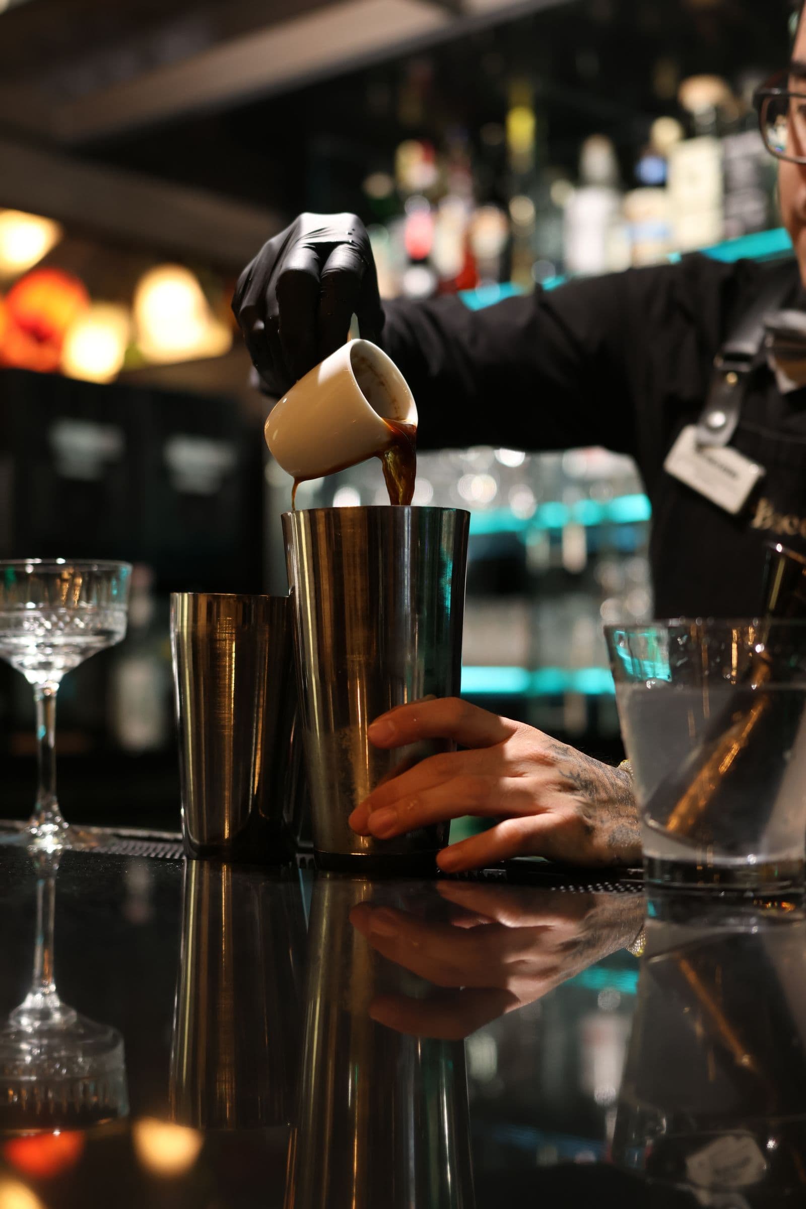 Cận cảnh tay bartender rót espresso vào Boston shaker trên quầy bar tối — kỹ thuật và dụng cụ pha chế